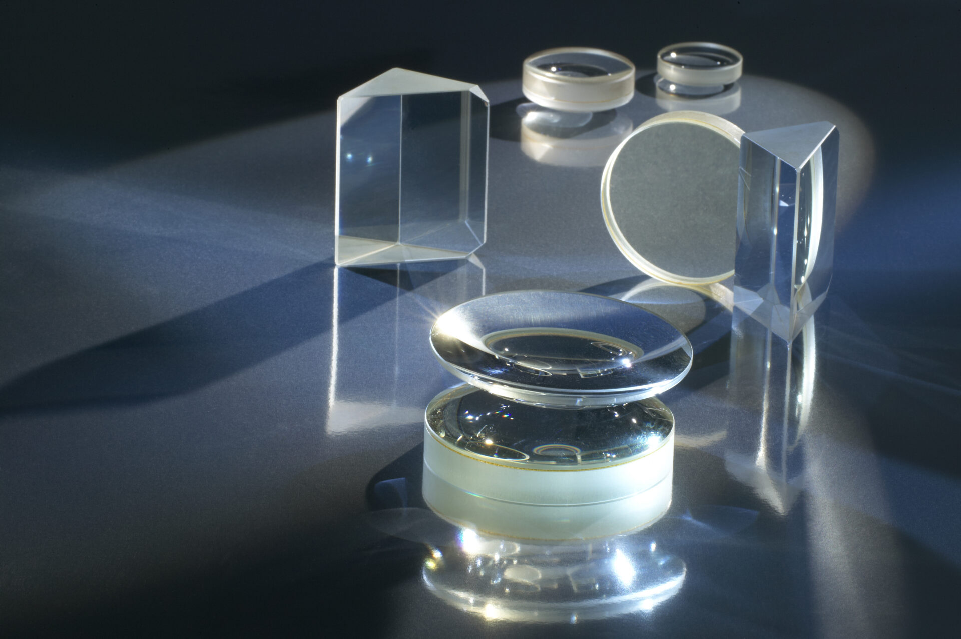 Optical glass lenses and prisms illuminated on a dark reflective surface.